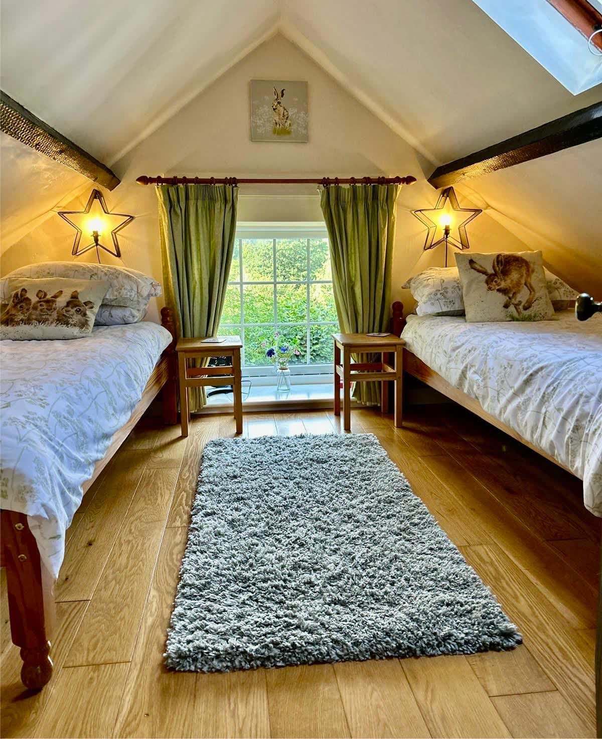 Cozy twin bedroom in a renovated milking parlour featuring two single beds, exposed wooden beams, and a green-curtained window.
